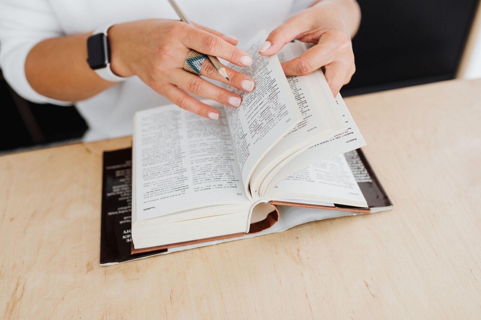 A person at a wooden desk leafing through a dictionary with a pencil in hand