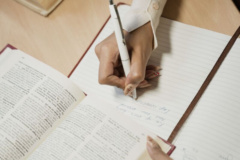 A hand writing English phrases into a notebook next to an open dictionary